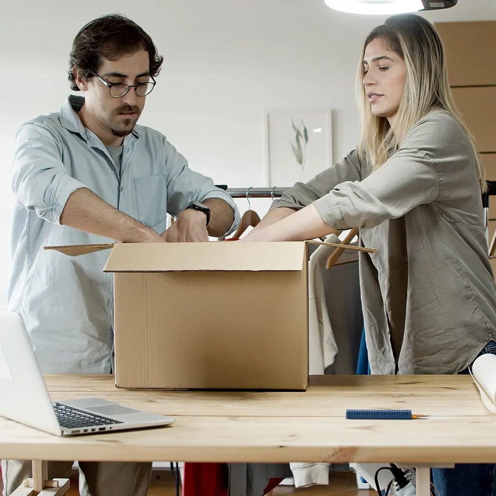 Two people packing a cardboard shipping box at a table with a laptop and packaging supplies.