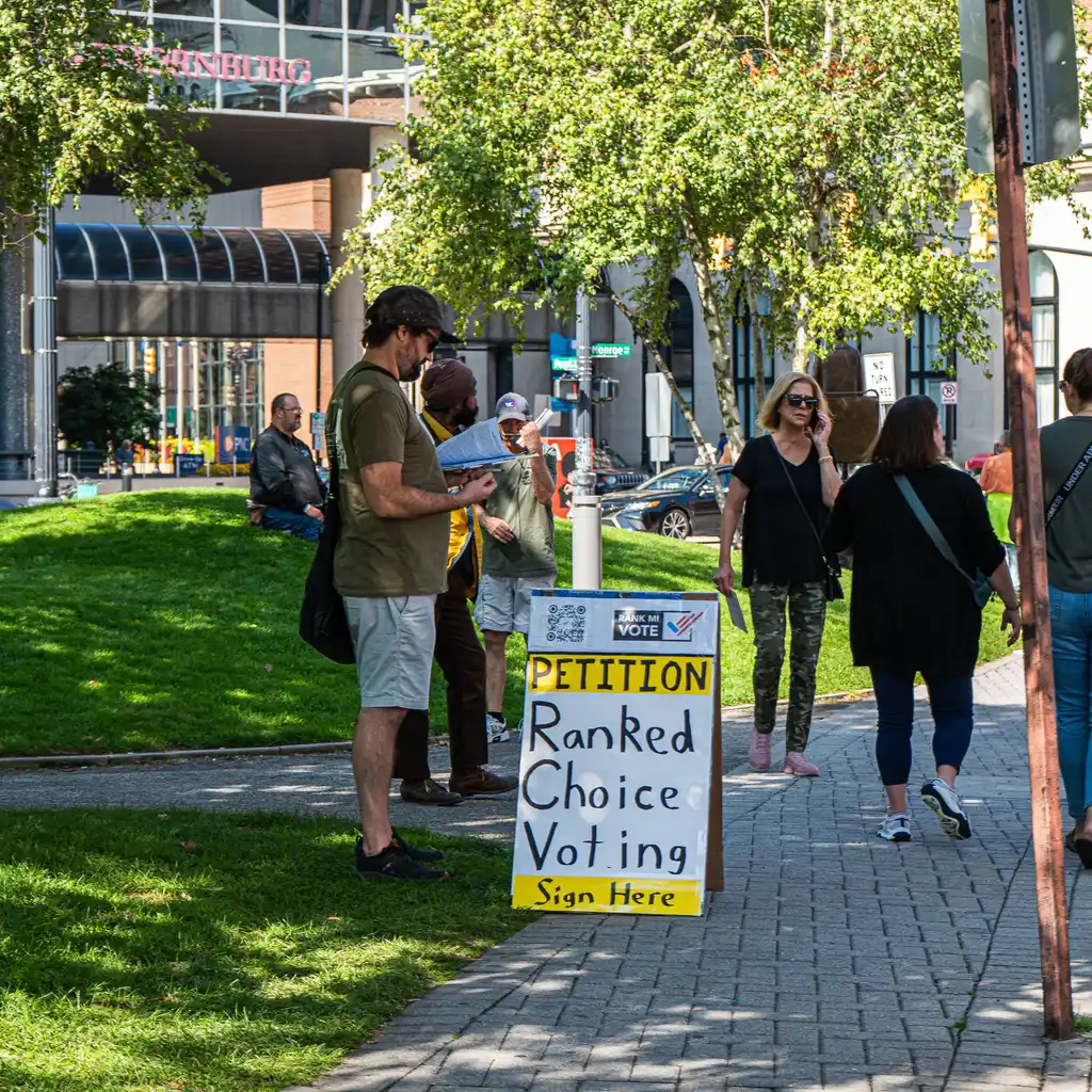 A canvasser speaking with passersby beside a petition sign.