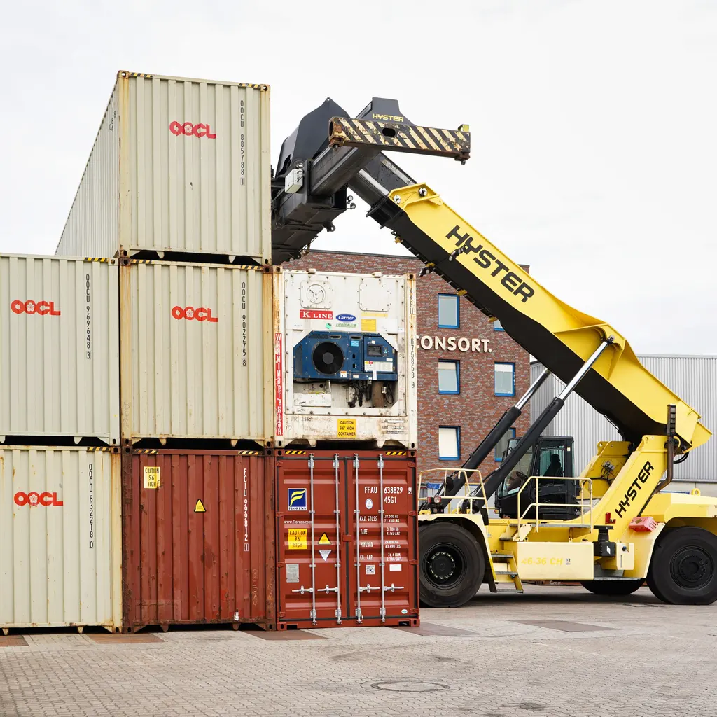 A reach stacker handling a shipping container beside stacked containers.