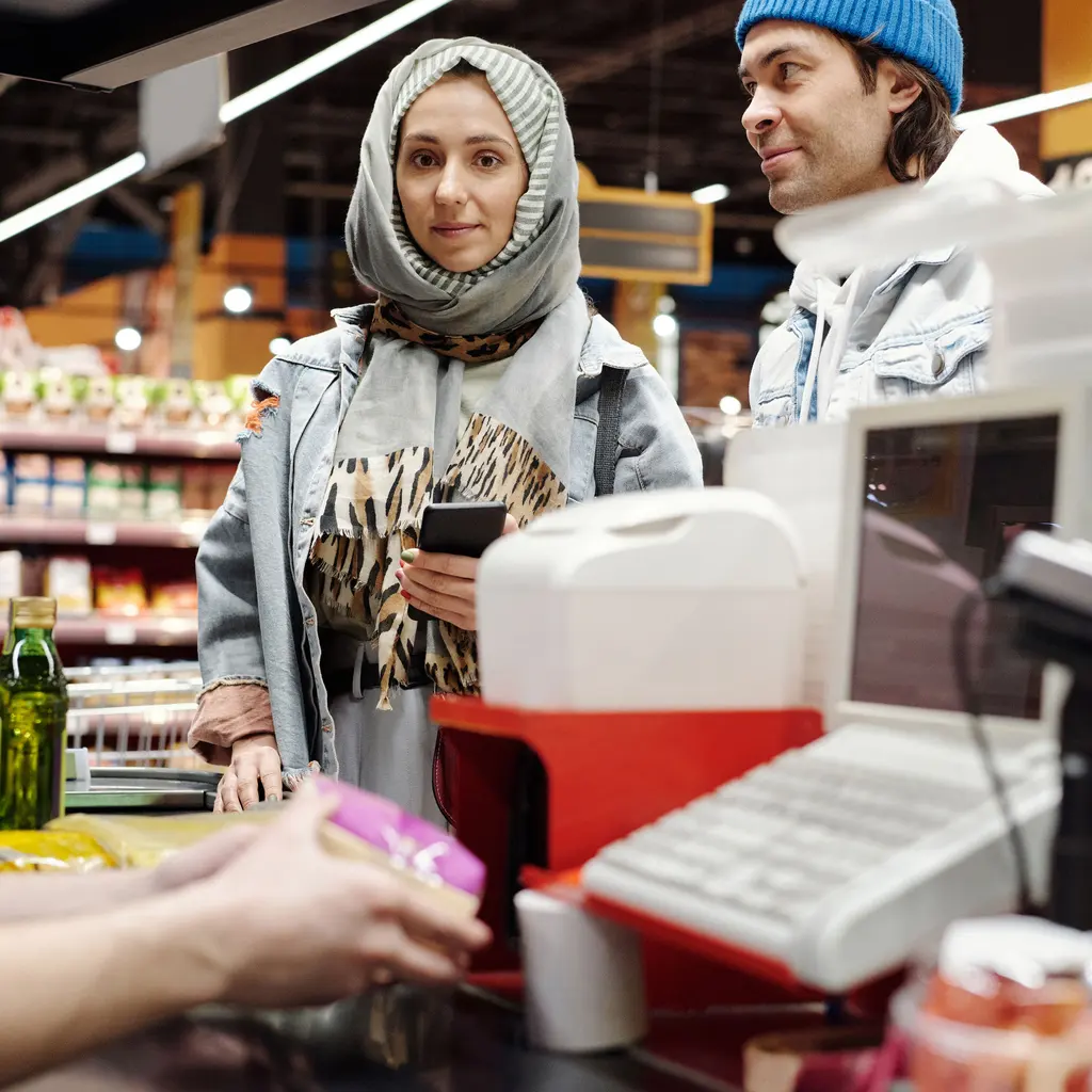 Customers waiting beside groceries and checkout equipment at a supermarket counter.