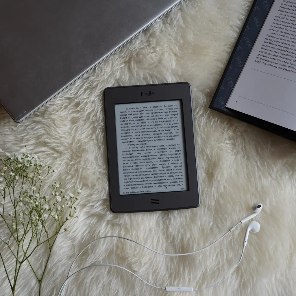 An e-reader displaying text beside a paper book, laptop and earbuds.