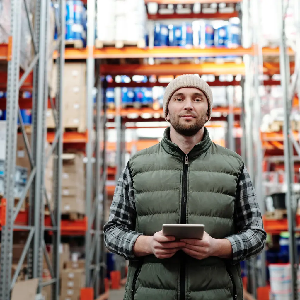A warehouse worker holding a tablet beside stocked pallet racks.