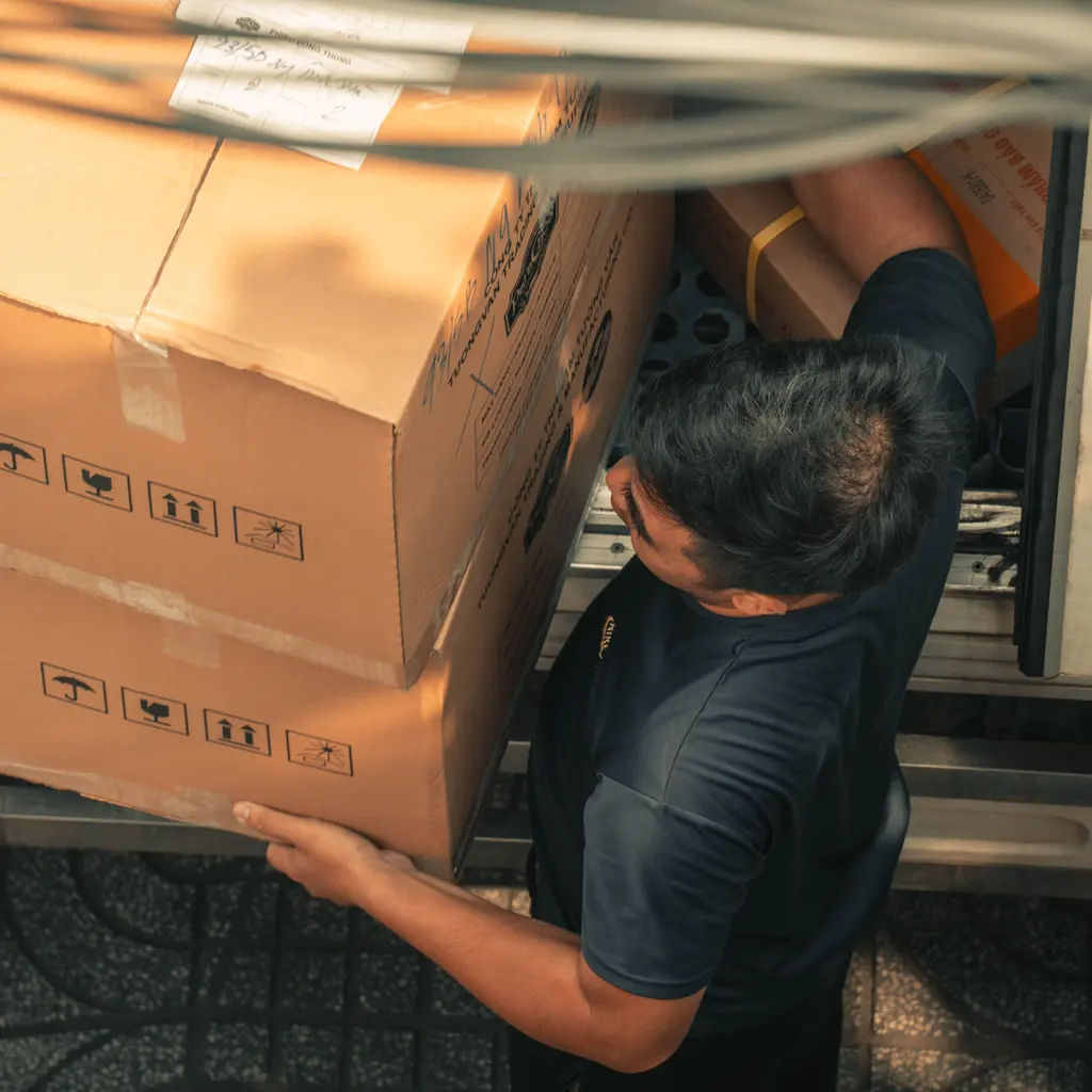 A delivery worker moving stacked cardboard boxes at the back of a truck.
