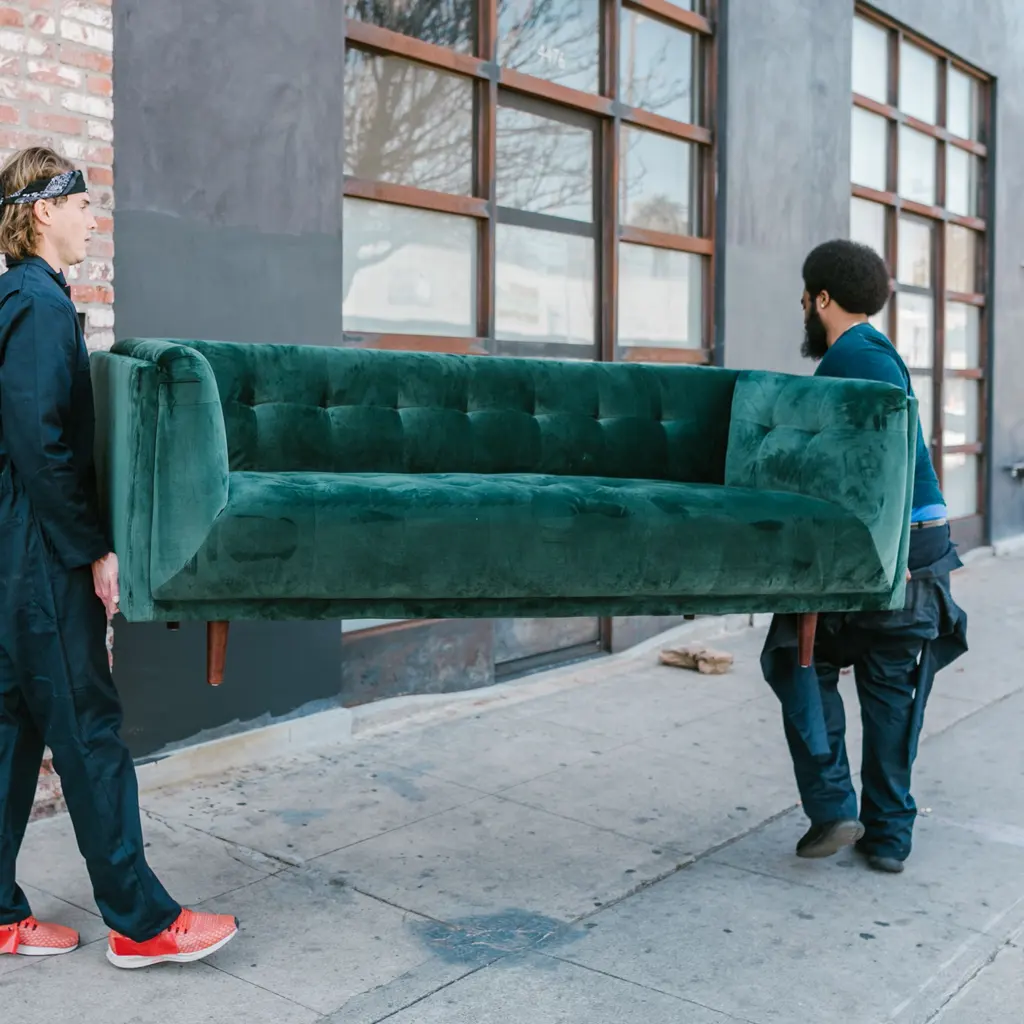 Two people carrying a green upholstered sofa outside a building.