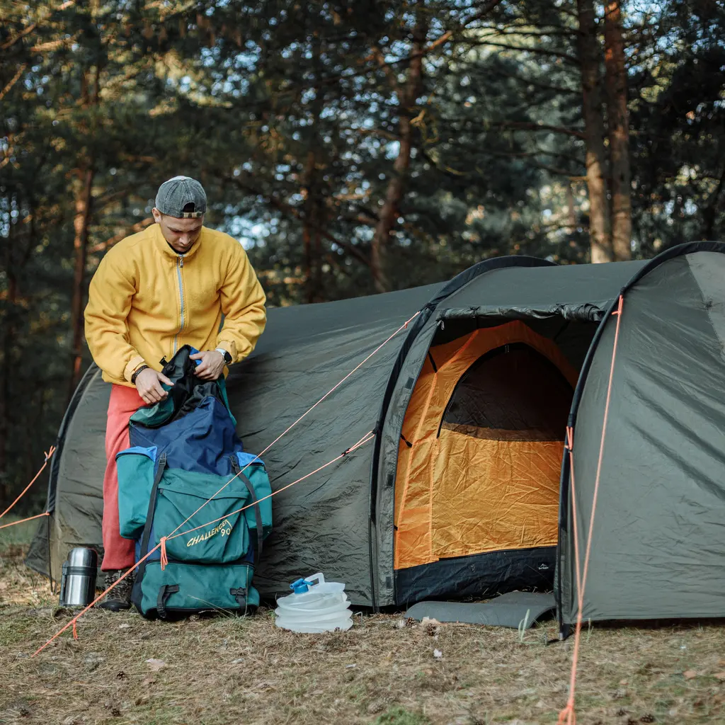 A camper arranging a large backpack beside a green tent in woodland.