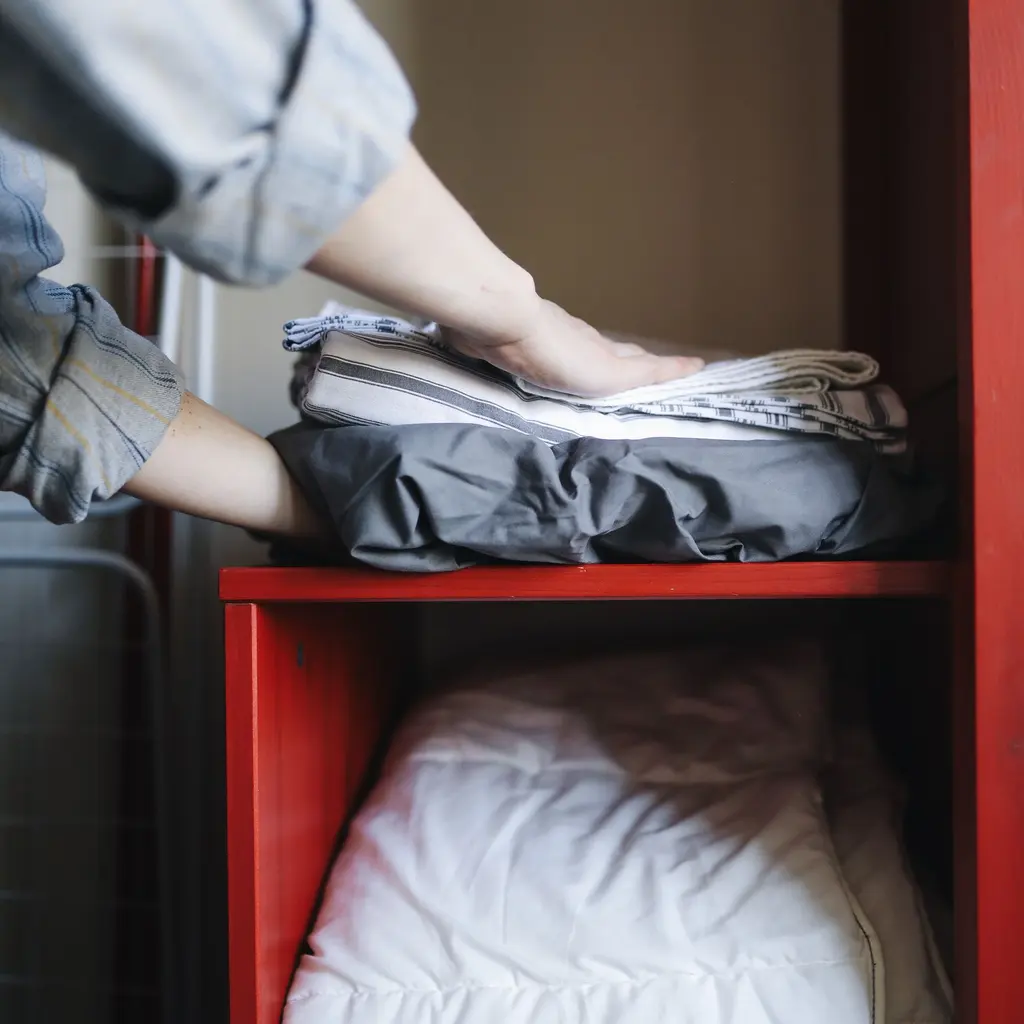 Hands folding clothing into a storage space on a household shelf.