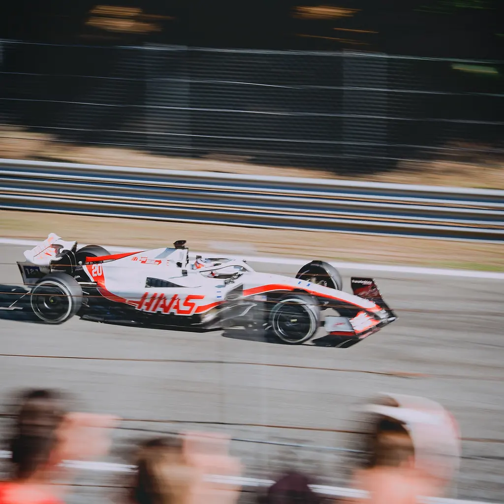 A racing car passing spectators and safety barriers on a motor-racing circuit.