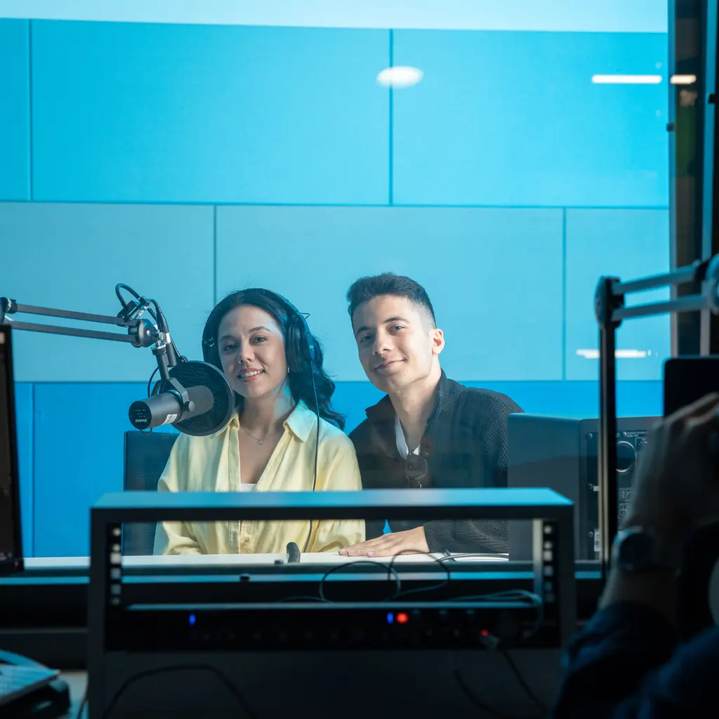 Two broadcasters at microphones behind the glass of a radio studio.