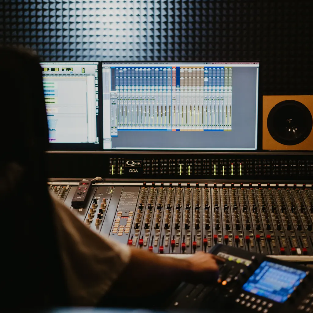 An engineer working at an audio mixing console beneath recording-software displays.