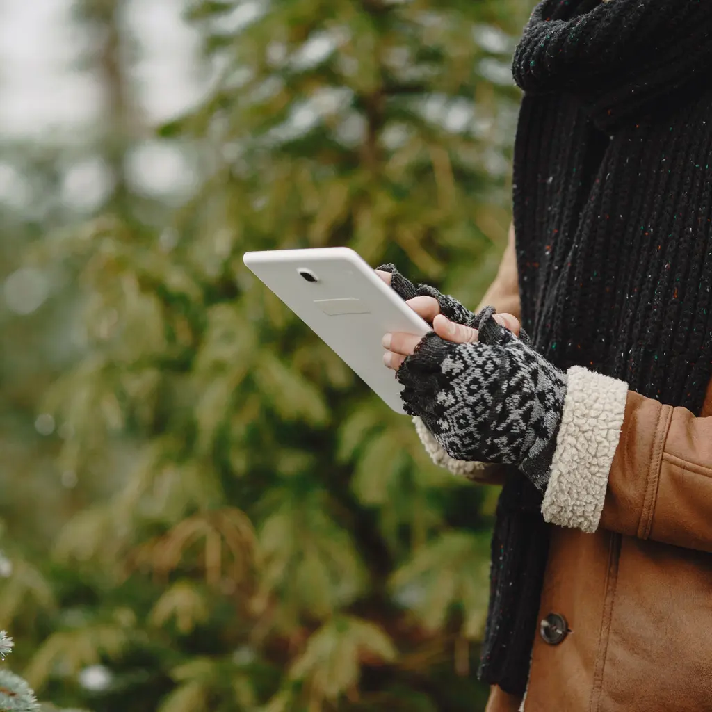 Gloved hands using a tablet beside snow-dusted evergreen trees.