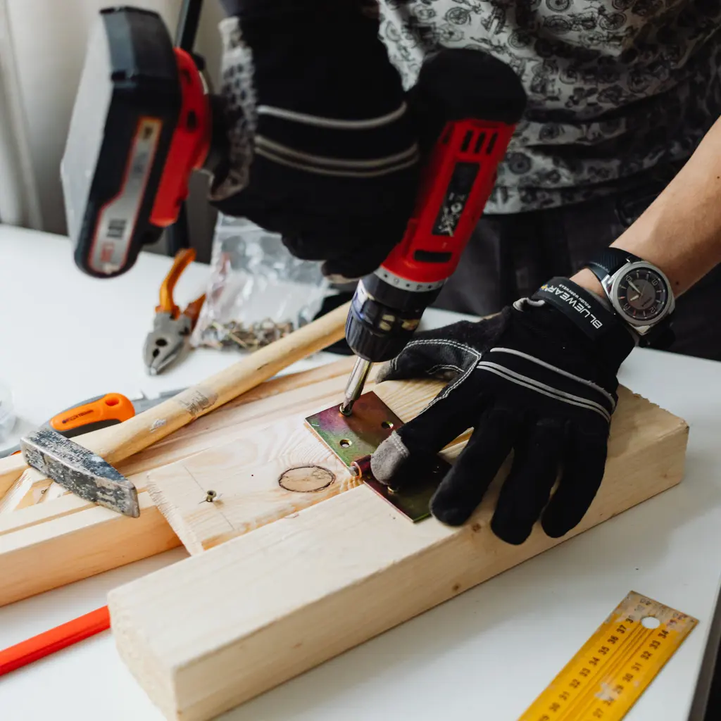 Gloved hands using a red power drill on a wooden workpiece.